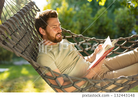 Caucasian bearded young man lying in hammock with a book in hands Caucasian bearded young man lying in hammock with a book in hands 132443148