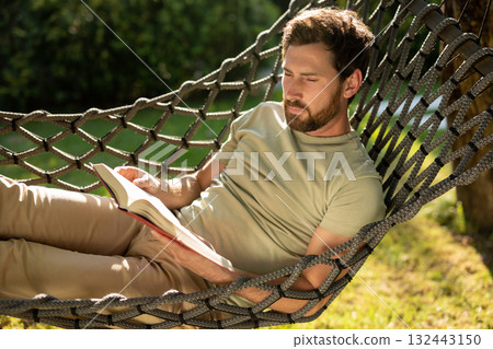 Caucasian bearded young man lying in hammock with a book in hands Caucasian bearded young man lying in hammock with a book in hands 132443150