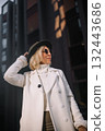 Stylish woman in a white coat and hat poses confidently outdoors against a modern urban backdrop 132443686