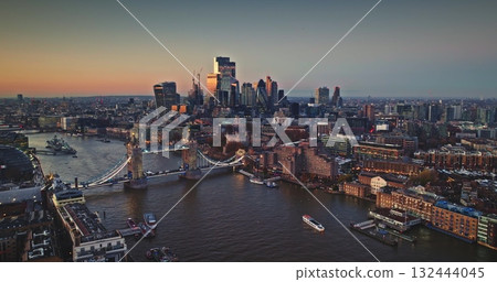 UK, London cityscape at sunset, iconic Tower Bridge spanning the River Thames, modern skyscrapers reflecting golden light against the twilight sky. Travel destination. Aerial drone flight panorama 132444045