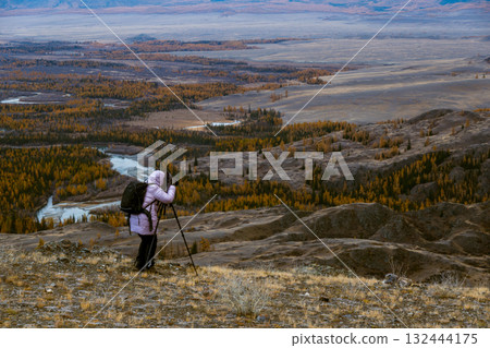 Photographer with tripod capturing expansive autumn landscape from hill overlooking river and forest 132444175