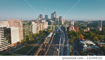Sydney skyline city architecture, buildings and busy infrastructure, multi-lane highway and train lines in warm morning light under a clear blue sky. Australia modern cityscape. Aerial drone footage 132444181