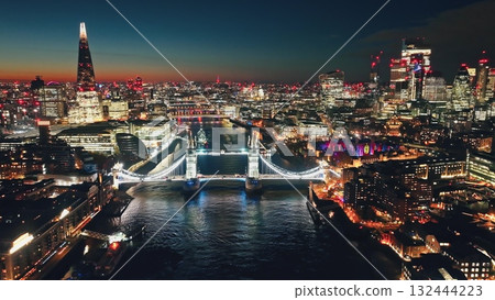 UK, London: Aerial view of famous Tower Bridge at night, The Shard and other landmarks along the Thames River, creating a stunning urban cityscape in colorful evening lights. Drone flight panorama 132444223