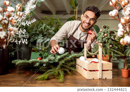 Man working in a flower shop and looking busy while decorating the christmas tree Man working in a flower shop and looking busy while decorating the christmas tree 132444821