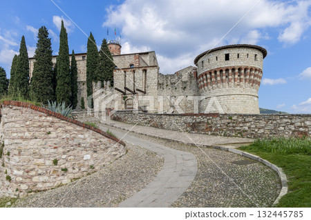 Brescia Castle rises beyond curving path with solid tower old bridge and calm trees around walls 132445785