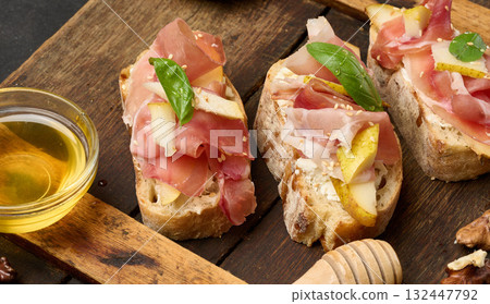 An arrangement of bruschetta with prosciutto, pear and cheese, served on a wooden board. The dish is garnished with drops of honey and basil leaves An arrangement of bruschetta with prosciutto, pear and cheese, served on a wooden board. The dish is garnished with drops of honey and basil leaves 132447792
