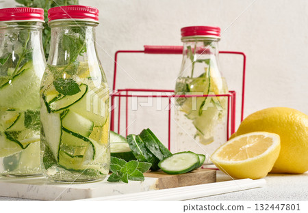 Glass bottles with red lids, filled with detox water containing cucumber and lemon slices and mint leaves, white table 132447801