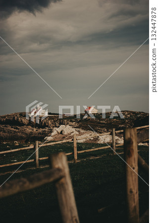 Coastal homes under a dramatic sky. Two houses sit atop a rocky hill, framed by a wooden fence and a moody sky. The scene evokes a sense of tranquility and isolation. Rural landscape scene. Cantabria. 132447878