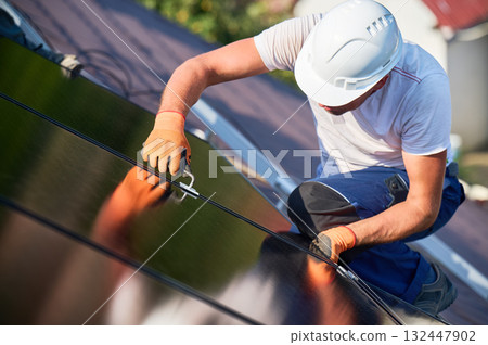 Worker building photovoltaic solar panel system on rooftop of house. Close up of man engineer in helmets and gloves installing solar module with help of hex key outdoors. Alternative energy. Worker building photovoltaic solar panel system on rooftop of house. Close up of man engineer in helmets and gloves installing solar module with help of hex key outdoors. Alternative energy. 132447902
