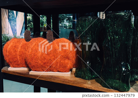 Felted orange pumpkins and green textured glass vases displayed on a wooden retail shelf. Thanksgiving decor, soft sculpture design, fall display styling, natural material accents Felted orange pumpkins and green textured glass vases displayed on a wooden retail shelf. Thanksgiving decor, soft sculpture design, fall display styling, natural material accents 132447979