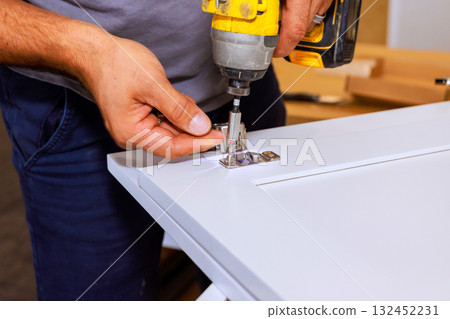 Installer uses screwdriver drill to attach hinges to cabinet door inside 132452231