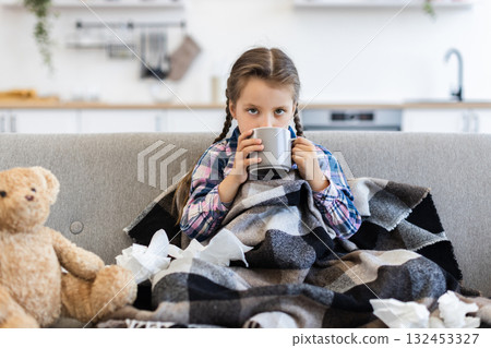 A young girl, wrapped in a blanket, sits on a couch, holding a mug. Tissues and a teddy bear are nearby, suggesting illness. 132453327