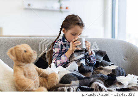 A child, possibly feeling unwell, is seen indoors, wrapped in a blanket, and holding a mug. A teddy bear and tissues are nearby. 132453328