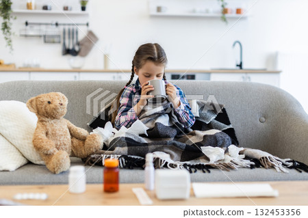 A young girl, wrapped in a blanket, sits on a couch and drinks from a mug, surrounded by medicine and tissues, looking unwell. A young girl, wrapped in a blanket, sits on a couch and drinks from a mug, surrounded by medicine and tissues, looking unwell. 132453356