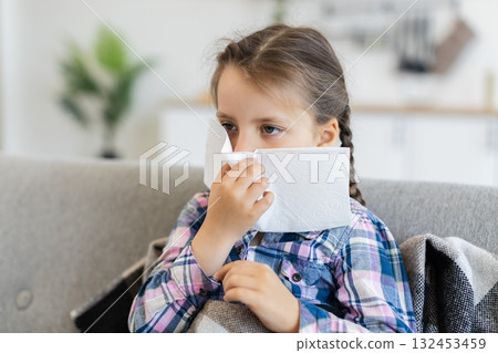 A young girl with braids is sitting on a couch, holding a tissue to her nose, possibly suffering from a cold or allergies. A young girl with braids is sitting on a couch, holding a tissue to her nose, possibly suffering from a cold or allergies. 132453459