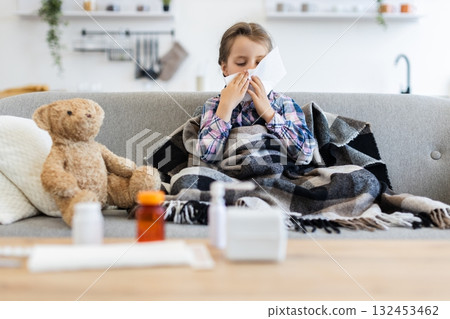 A young girl, wrapped in a blanket, is blowing her nose with a tissue while sitting on a couch. A teddy bear and medicine are nearby. 132453462