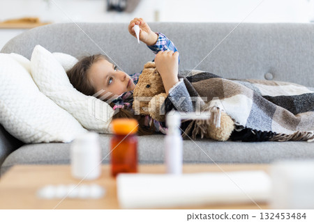 A young girl, wrapped in a blanket, looks at a thermometer while resting on a sofa. Medicine bottles are on the table. 132453484