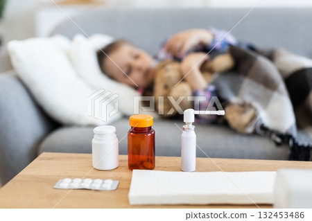 A young child rests on a sofa, appearing unwell, with various medications placed on a table in the foreground, suggesting illness. 132453486