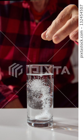 Older man dropping tablet into water glass creating dissolving bubbles Older man dropping tablet into water glass creating dissolving bubbles 132454507