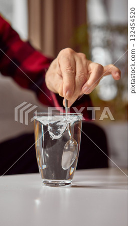 Hand stirring clear water in glass with metal spoon on white table Hand stirring clear water in glass with metal spoon on white table 132454520