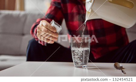 Senior man pouring hot water into glass while preparing powdered medicine drink Senior man pouring hot water into glass while preparing powdered medicine drink 132454521
