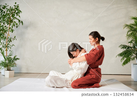 Masseuse making thai yoga massage. Woman therapist doing traditional massage treatment, stretching female patient's body. Client sitting on mat, enjoying hand massage in calm atmosphere. 132458634