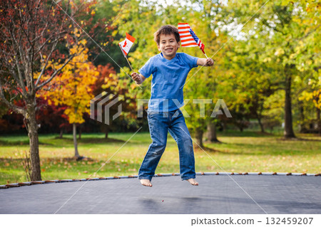 Cheerful boy jumps barefoot on a trampoline holding USA and Canada flags in a colorful autumn park Cheerful boy jumps barefoot on a trampoline holding USA and Canada flags in a colorful autumn park 132459207