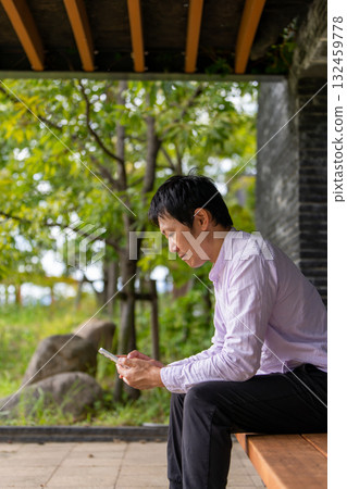 一位商人坐在公園長椅上查看通知,一位男士坐在綠樹環繞的長椅上專注於智慧型手機 一位商人坐在公園長椅上查看通知,一位男士坐在綠樹環繞的長椅上專注於智慧型手機 132459778