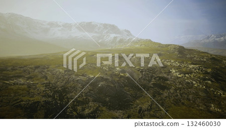 Rolling green hills stretch toward majestic snow capped mountains in the distance. A soft morning light bathes the landscape in a calm, tranquil atmosphere. 132460030