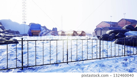 Snow blankets the ground at a secluded research station, with colorful structures peeking through the icy landscape. Tall rocks stand around, bathed in a serene winter light. 132460040