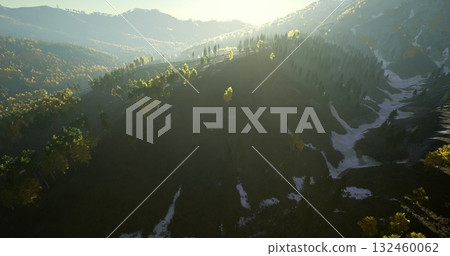 Gentle sunlight cascades over rugged mountains, highlighting lush greenery and patches of snow. Nature awakens as morning light bathes the landscape in a warm glow, creating tranquility. 132460062