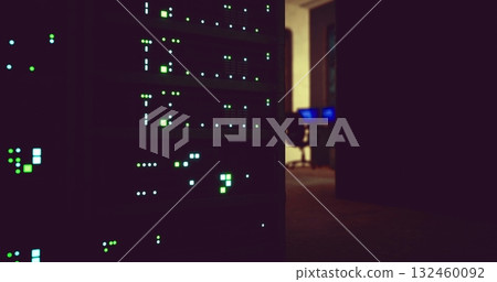 In a dimly lit office, multiple data servers glow with green and blue lights. The setting suggests late hours as a work desk with computers sits in the background, indicating active monitoring. 132460092