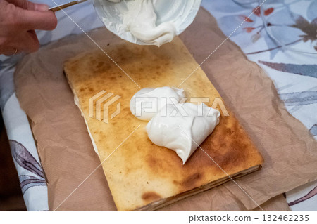 Close-up of a hand adding creamy sauce onto a freshly baked flatbread, showcasing the culinary process and textures in a cozy kitchen setting with natural lighting Close-up of a hand adding creamy sauce onto a freshly baked flatbread, showcasing the culinary process and textures in a cozy kitchen setting with natural lighting 132462235