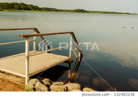 Tranquil lakeside scene featuring wooden dock extending over calm water, surrounded by lush greenery and distant trees, creating a serene natural atmosphere for relaxation 132462236