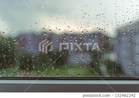 Rain droplets on a windowpane create a blurred view of a garden and buildings outside, capturing the essence of a rainy day atmosphere and nature's beauty 132462242