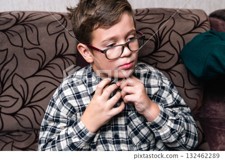 Young boy with glasses adjusting his shirt collar while sitting on a patterned couch, showcasing a moment of thoughtful preparation for a special occasion 132462289
