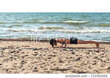 Individual performing plank on sandy beach, Person maintaining strict plank position amid tranquil seaside environment Individual performing plank on sandy beach, Person maintaining strict plank position amid tranquil seaside environment 132462608