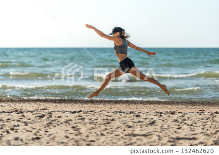 Woman jumps freely over shore, Energetic performance showcasing flexibility and strength against seaside horizon Woman jumps freely over shore, Energetic performance showcasing flexibility and strength against seaside horizon 132462620