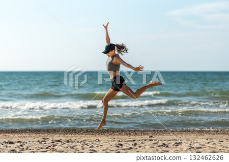 Dynamic female athlete on sunny shoreline, Cheerful woman in energetic motion leaping at coast with windblown hair 132462626