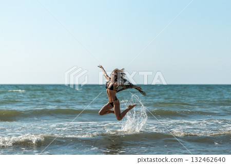 Joyful seaside leap, Woman enthusiastically leaping through mild coastal waters with uplifted arms and cheerful spirit 132462640