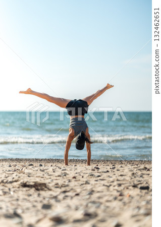 Joyful female athlete performing acrobatic move at coast, Playful lady showcasing athleticism with cartwheel by ocean 132462651