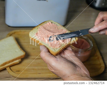 Medium view of a hand holding a slice of bread and spreading creamy spread with a knife. Medium view of a hand holding a slice of bread and spreading creamy spread with a knife. 132462714