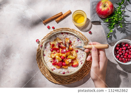 Hand holding spoon in to plate of oatmeal with apple, cranberry and cinnamon on a light background 132462862