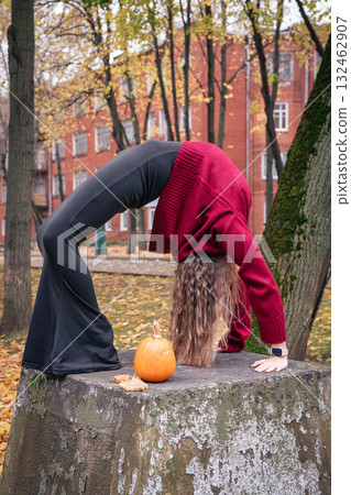 Teen girl in a red sweater performs a yoga backbend on a stone surface beside a small pumpkin in an autumn park near Timiryazev Academy Teen girl in a red sweater performs a yoga backbend on a stone surface beside a small pumpkin in an autumn park near Timiryazev Academy 132462907