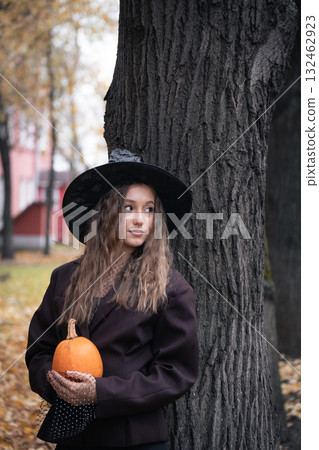 Teen girl in witch hat holding a pumpkin near a tree, surrounded by autumn leaves and soft natural light, creating a mysterious Halloween mood 132462923