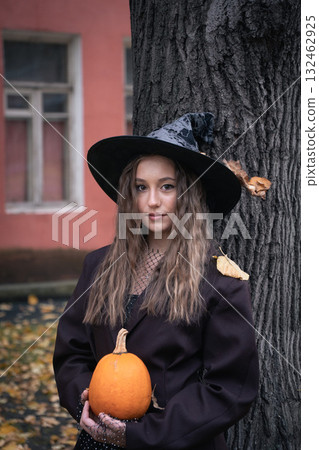 Teen girl in witch hat holding a pumpkin near a tree, surrounded by autumn leaves and soft natural light, creating a mysterious Halloween mood 132462925