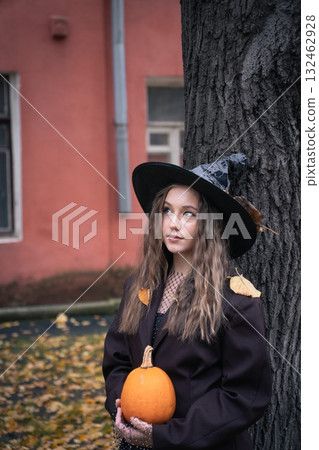 Teen girl in witch hat holding a pumpkin near a tree, surrounded by autumn leaves and soft natural light, creating a mysterious Halloween mood 132462928