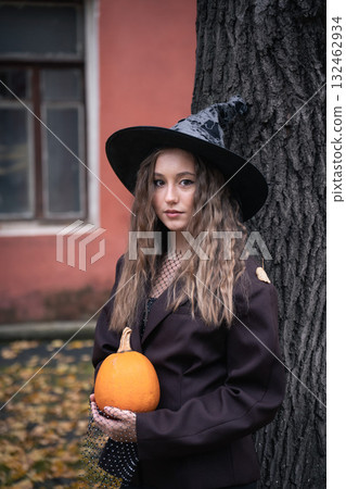 Teen girl in witch hat holding a pumpkin near a tree, surrounded by autumn leaves and soft natural light, creating a mysterious Halloween mood 132462934