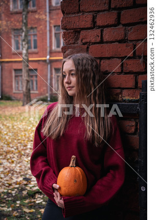 Teen girl in a red sweater holds a pumpkin while leaning against an old brick wall with autumn leaves on the ground in the Timiryazevs Academy, Moscow Teen girl in a red sweater holds a pumpkin while leaning against an old brick wall with autumn leaves on the ground in the Timiryazevs Academy, Moscow 132462936