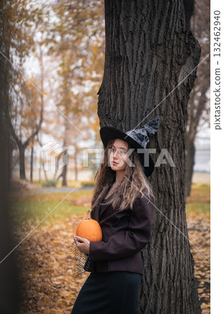 Teen girl in witch hat holding a pumpkin near a tree, surrounded by autumn leaves and soft natural light, creating a mysterious Halloween mood 132462940